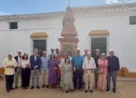 Grupo de personas posando en el jardín frente a un monumento con estatua de la Virgen vestida de Pastora y edificio blanco con ventanas con rejas verdes y toldos amarillos