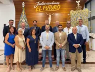 Grupo de trece personas posando en interior con dos estandartes religiosos y un crucifijo sobre fondo de madera con texto Juntos escribimos el futuro