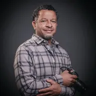Hombre de mediana edad con camisa de cuadros sostiene una cámara fotográfica y sonríe frente a fondo gris oscuro