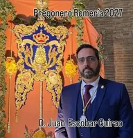 Hombre con traje azul y corbata roja posa junto a un estandarte rociero azul y dorado con corona y flores amarillas en fondo naranja