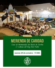Gran grupo de personas sentadas en largas mesas en un patio decorado con luces y banderines durante una merienda solidaria nocturna en la Casa de Hermandad del Rocío en Sevilla