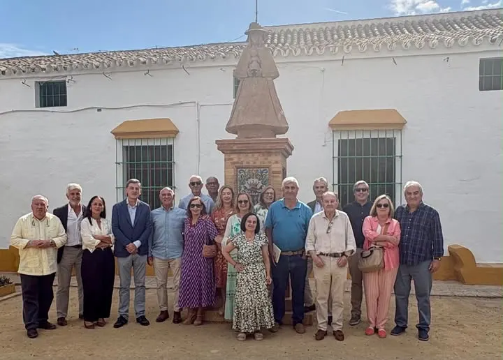 Grupo de personas posando en el jardín frente a un monumento con estatua de la Virgen vestida de Pastora y edificio blanco con ventanas con rejas verdes y toldos amarillos