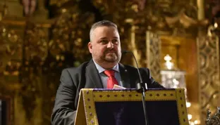 Hombre con traje oscuro y corbata roja habla en un atril decorado con tela morada y dorada en un interior con ornamentos dorados religiosos