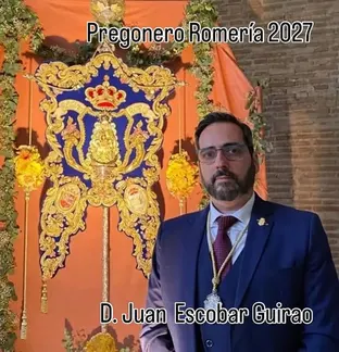Hombre con traje azul y corbata roja posa junto a un estandarte rociero azul y dorado con corona y flores amarillas en fondo naranja