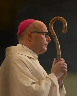 Hombre con vestimenta clerical blanca y solideo rojo sostiene un báculo pastoral mirando hacia la derecha sobre fondo oscuro