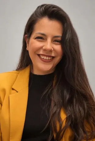 Mujer joven sonriente con cabello largo y oscuro, lleva chaqueta mostaza y camiseta negra, posando frente a fondo gris claro