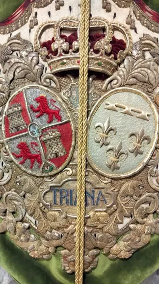 Bordado detallado en tonos dorados y rojos con escudos, corona real y la palabra Triana sobre fondo verde en estandarte religioso